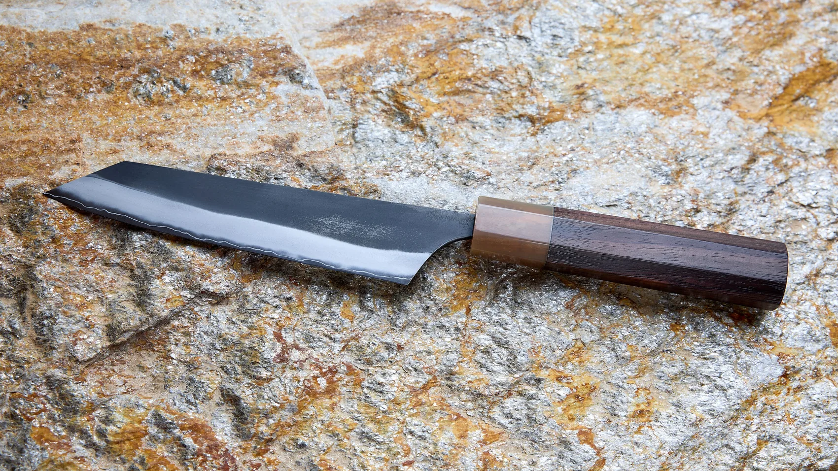 A Japanese chef's knife with a black forged blade and an octagonal wooden handle lies on gold-flecked granite