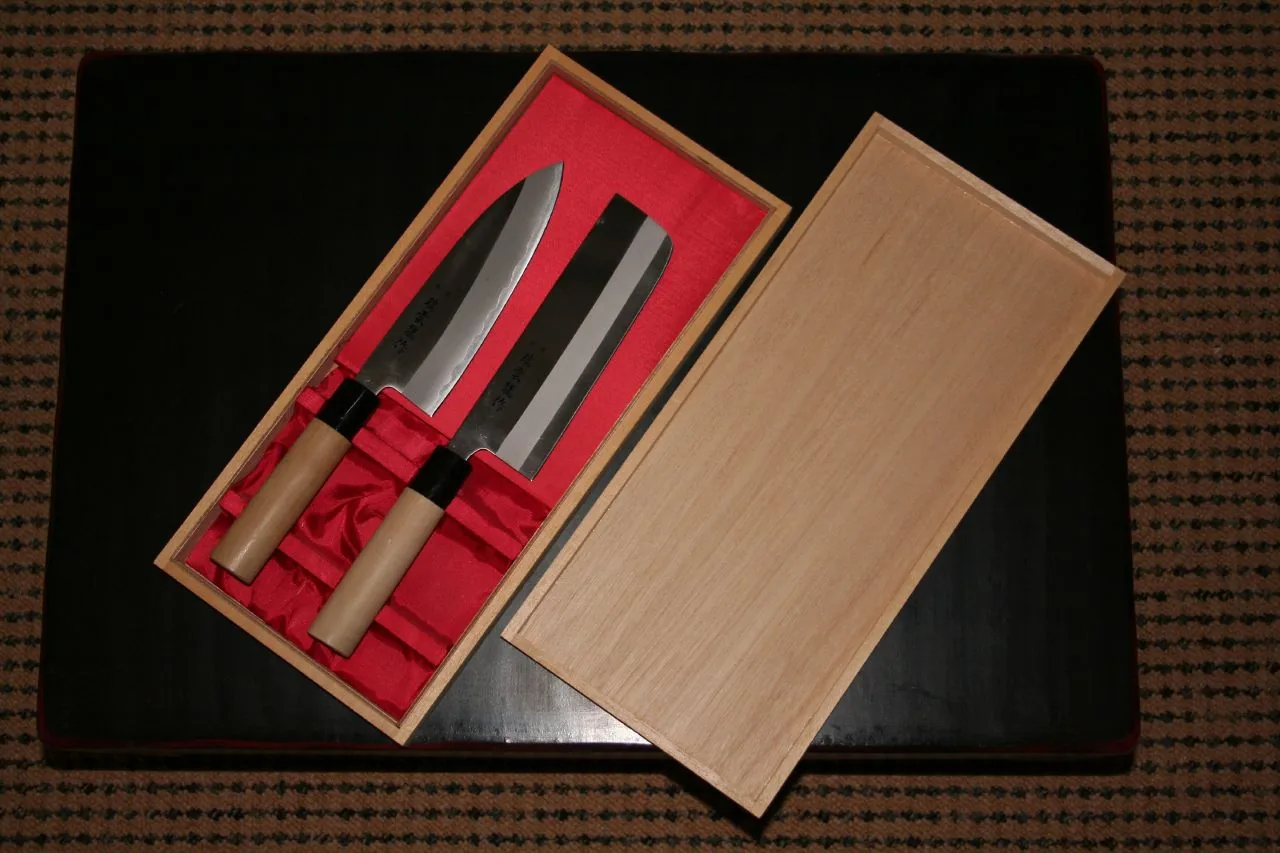 Two Japanese kitchen knives with pale wooden handles lie in an open wooden gift box lined with red silk
