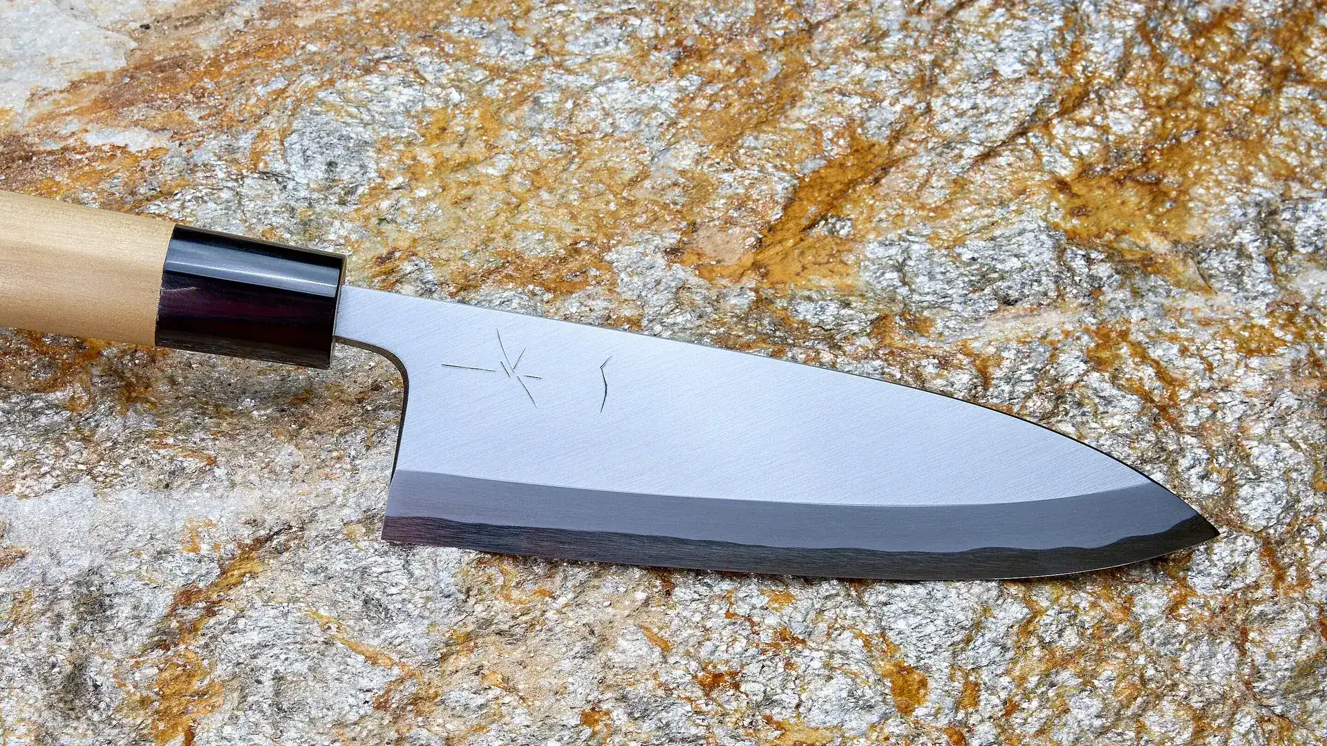 A Japanese deba kitchen knife with a mirror-polished blade and a dark forged edge lies on gold-flecked granite
