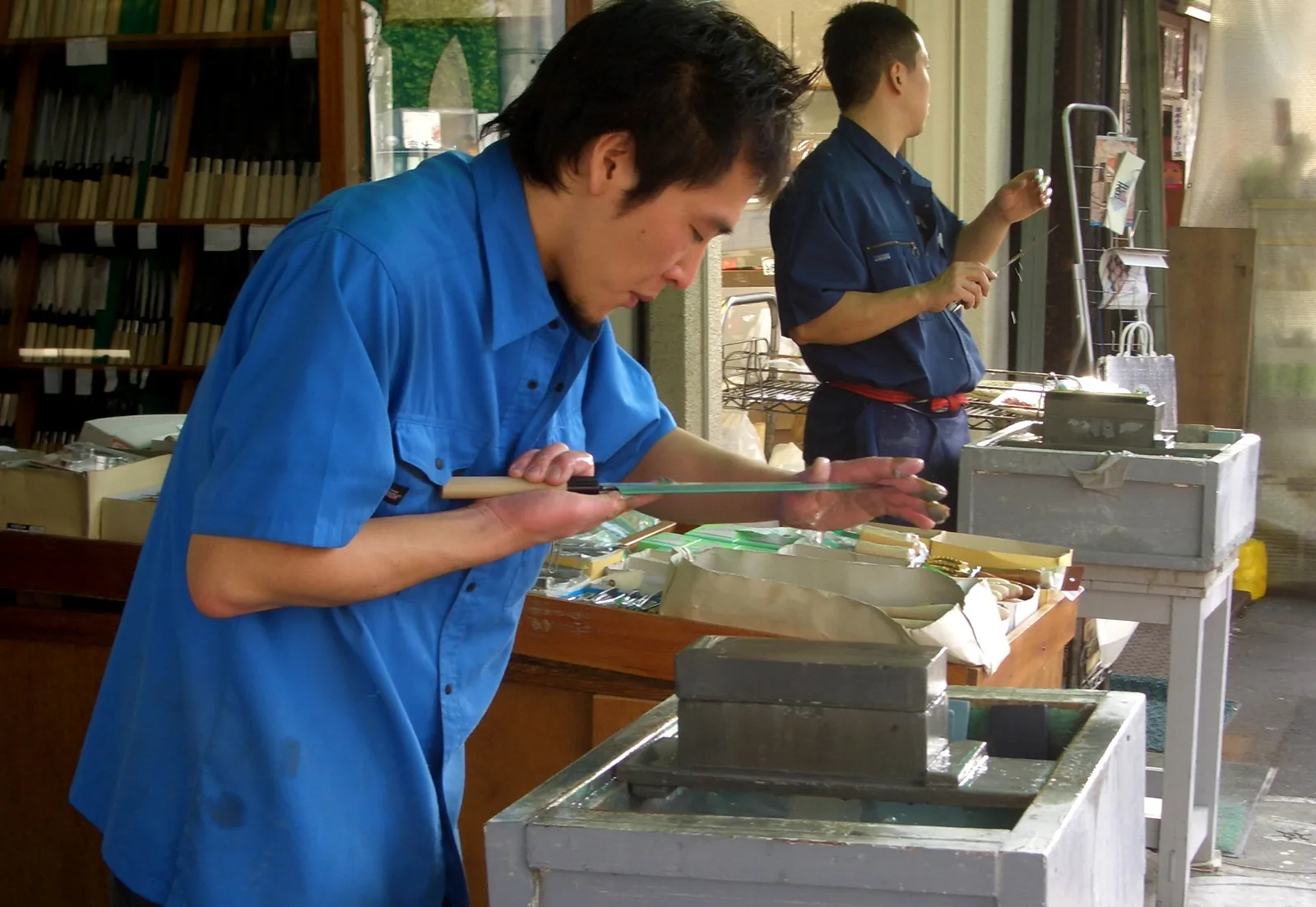 A knife sharpener in a blue shirt leans over a blade beside wooden water-stone tanks at a Tokyo knife stall