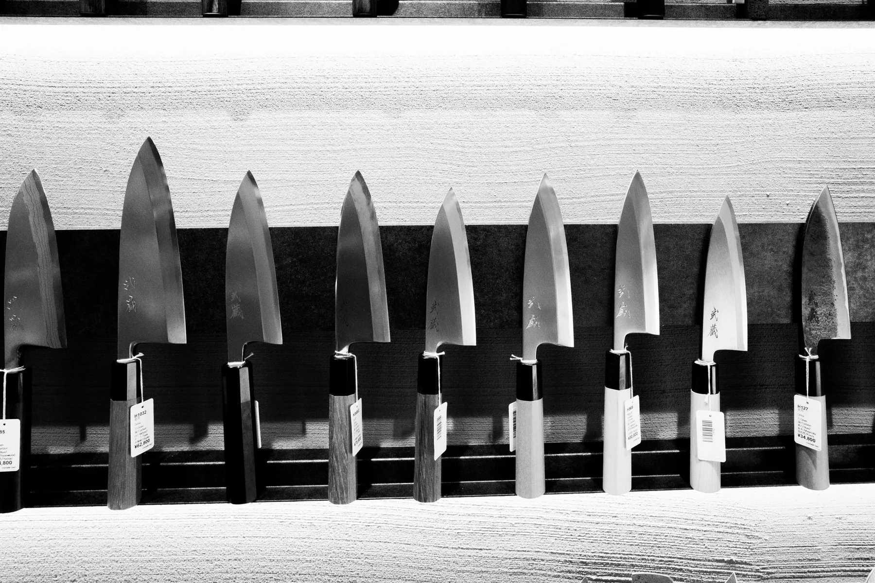 Black-and-white photograph of a row of Japanese deba knives with wooden handles hanging blade-up on a rack
