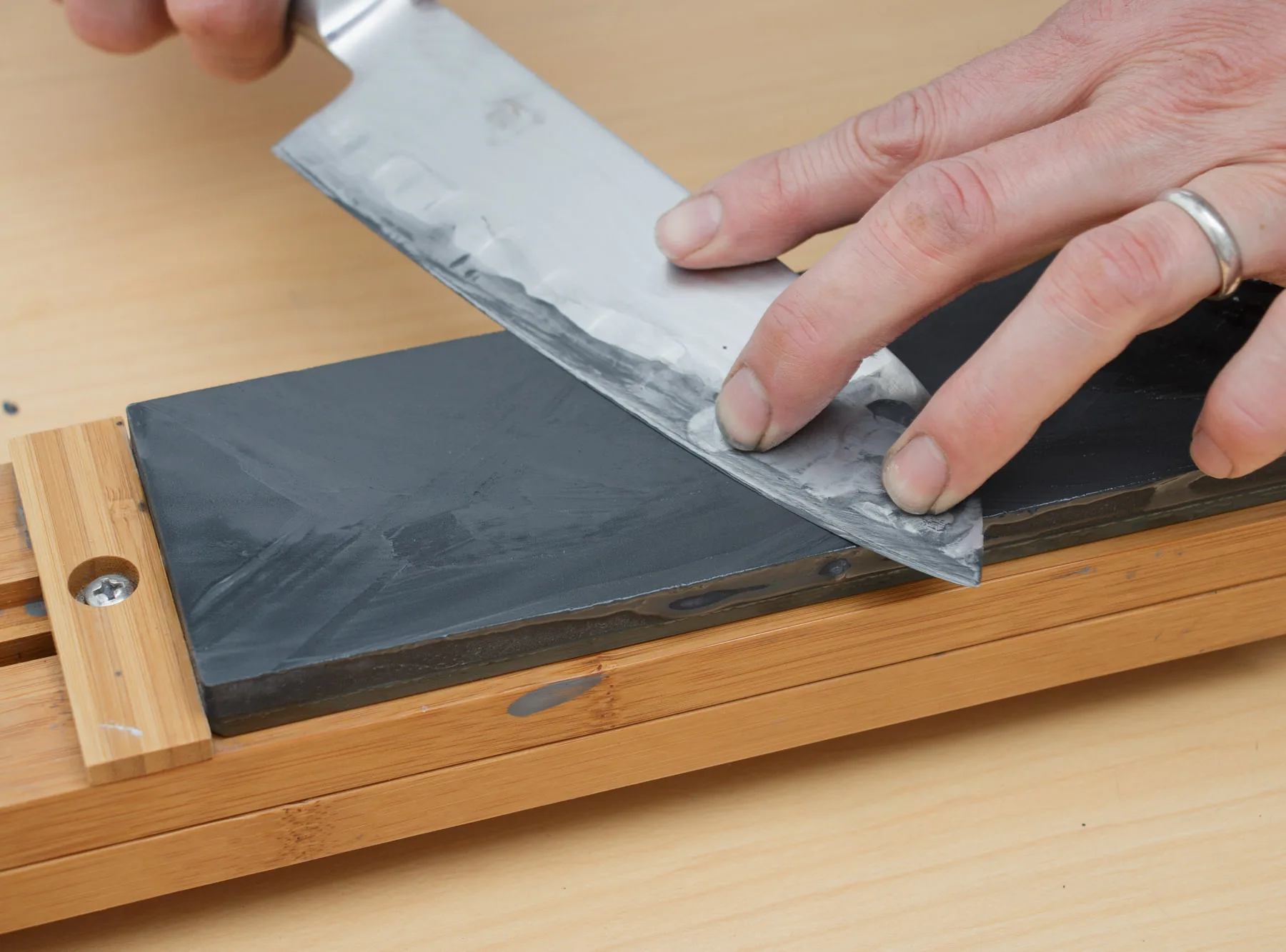 A hand presses the flat of a knife against a dark Japanese water stone set in a wooden holder